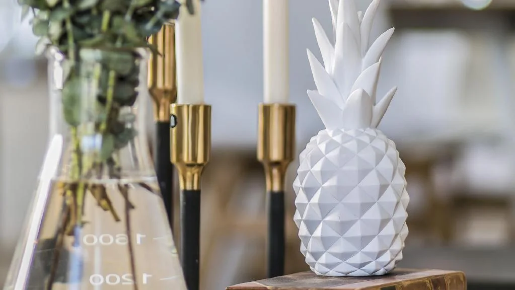 Modern home decor featuring a white pineapple sculpture, gold candle holders, and flowers in a beaker vase. Interior design trends.